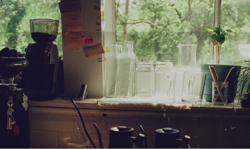 A back counter with coffee brewing equipment and cups with a view of  green trees out the window in the back. 