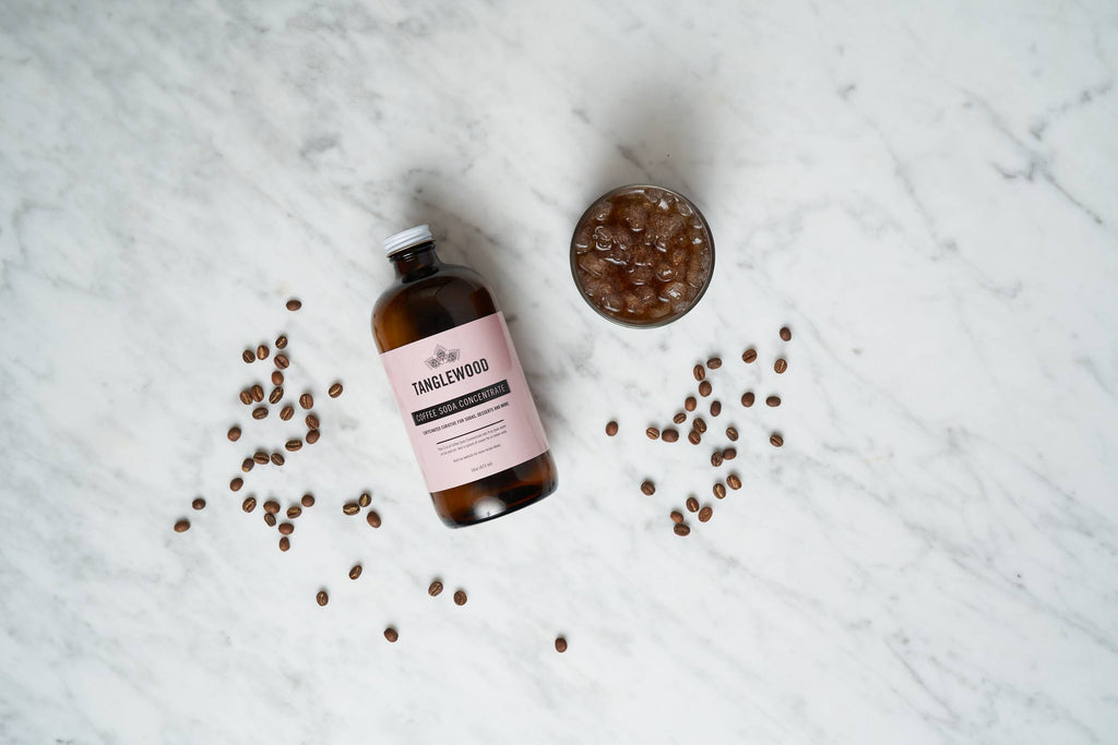Tanglewood Coffee Soda Concentrate bottle next to prepared beverage surrounded by whole coffee beans on a marble table.